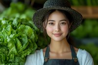 Young Asian female farmer wearing straw hat and apron standing among fresh green lettuce plants in vegetable garden, suitable for agriculture and farming content.