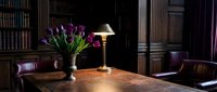 Elegant dark wood library interior with purple tulips and glowing brass lamp on antique desk, leather chairs, bookshelves in background.