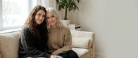 Two young women friends embracing on beige sofa in bright living room with natural light. Cozy home interior friendship bonding moment for lifestyle content.