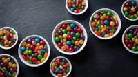 Colorful candy-coated chocolates in white bowls arranged on dark slate surface. Sweet treats for parties, celebrations, and dessert displays.
