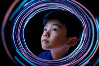 Young Asian boy gazing upward through glowing neon circular light rings in pink and blue colors against dark background, creating futuristic portrait with dramatic lighting effect.