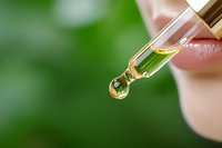 Close-up of dropper with golden CBD oil or essential oil drop against blurred green natural background, wellness and natural medicine concept.