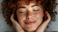 Young woman with natural freckles, curly red hair, and closed eyes expressing serenity and joy while holding face in hands against white background. Wellness concept.