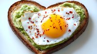 Heart-shaped toast with mashed avocado spread and sunny side up egg garnished with red pepper flakes on white background, healthy breakfast concept.