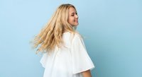 Young blonde woman with flowing hair in white shirt turning and smiling against blue background. Studio portrait for lifestyle and fashion concepts.