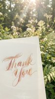 Thank you card with rose gold calligraphy lettering held outdoors against blurred garden background with blooming flowers and natural sunlight for gratitude and appreciation messaging.