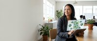 Young South Asian woman in business casual attire holding laptop with green sustainability icons in modern office workspace with plants and natural lighting.