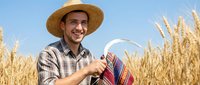Young Hispanic male farmer in straw hat and plaid shirt smiling while holding wicker basket in golden wheat field under blue sky for agriculture concepts.