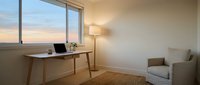 Modern home office workspace with wooden desk, laptop, and floor lamp beside large window overlooking sunset sky, minimalist interior design for remote work and productivity.