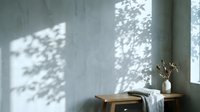 Minimalist interior with wooden table, white vase with dried branches, and natural light casting leaf shadows on gray wall.