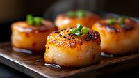 Seared sea scallops with caramelized golden-brown crust and green onion garnish on dark wooden serving board, close up view showing juicy texture and gourmet preparation.