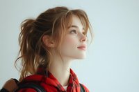 Young Caucasian woman with messy ponytail wearing red plaid shirt gazes upward with hopeful expression against light background, natural makeup and soft lighting.