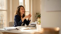 Happy businesswoman laughing while drinking coffee at office desk with laptop and documents in bright modern workspace with natural sunlight.