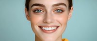 Young woman with bright white smile and natural makeup showing perfect dental care against blue background, expressing joy and confidence in close-up portrait.