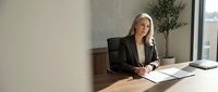 Mature woman with gray hair and light skin tone, wearing a black blazer, sitting at a wooden desk in a modern office environment, reviewing documents with a pen in hand.