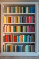 Colorful books arranged by spectrum on white wooden bookshelf, creating rainbow pattern across five shelves. Clean minimalist library organization for home decor.
