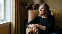 Senior Caucasian woman in black sweater sitting thoughtfully in vintage leather armchair by window, warm natural lighting creating peaceful retirement moment.