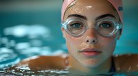Young female swimmer in pink cap and goggles emerges from turquoise pool water, closeup portrait showing determination and focus during training session.