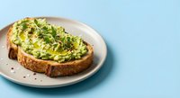 Fresh avocado toast on artisan bread with microgreens and seeds, served on ceramic plate against pastel blue background for healthy breakfast concepts.