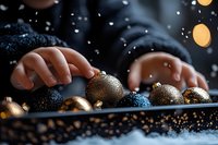 Child hands arranging elegant black and gold Christmas baubles on dark surface with glitter and bokeh lights, creating festive holiday atmosphere.