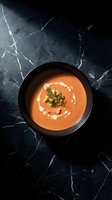 Creamy tomato soup with herbs and cream swirl in black bowl on dark marble surface for restaurant menu and food photography.