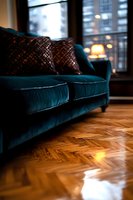 Luxurious teal velvet sofa with decorative pillows on herringbone hardwood floor, illuminated by warm window light in cozy modern living room interior.