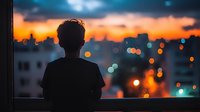 Young man silhouette against evening cityscape with blurred bokeh lights and dramatic orange sunset sky, viewed through window. Contemplative urban lifestyle scene.