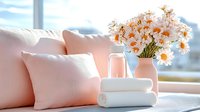 Fresh daisies in white vase with soft pink pillows and rolled towels on windowsill create serene spa atmosphere with natural morning light.