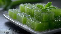 Green jelly cubes garnished with fresh mint leaves on metallic plate with water droplets, close up view showing translucent texture and glossy surface.