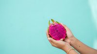 Hands holding vibrant pink dragon fruit with green crown against turquoise background, suitable for food, wellness, and tropical lifestyle content.