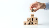 Hand placing wooden cube with target icon on stack of business blocks showing growth charts and dollar sign for goal achievement strategy.