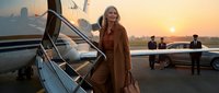 Elegant blonde businesswoman in luxury coat boarding private jet at sunset with professional crew and golden sky backdrop for corporate travel concepts.