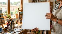 Female artist of mixed ethnicity holding blank white canvas in bright studio workspace with paintbrushes, art supplies, and windows overlooking landscape, ready for creative project.