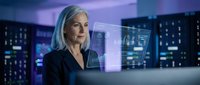 Mature woman with white hair wearing dark blazer works at computer terminal surrounded by holographic data displays and digital interface screens in modern tech environment.