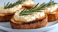 Toasted bread slices topped with creamy white spread and fresh rosemary sprigs on white plate. Appetizer canapes for party catering and restaurant menu.