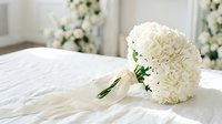White carnation bridal bouquet with satin ribbon on bed in elegant wedding suite with floral arrangements for romantic ceremony celebration.