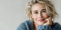 Smiling blonde Caucasian woman in cozy blue knit sweater with hand on chin, relaxed portrait on neutral background for lifestyle and wellness content.
