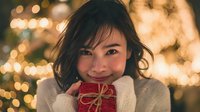 Young Asian woman holding small red gift box with twine bow against warm bokeh holiday lights background, creating festive Christmas atmosphere.