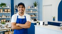 Young Hispanic male potter smiling with crossed arms in blue apron at ceramic studio workspace with shelves of pottery and plants in background.