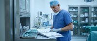 Male surgeon in blue scrubs reviewing medical documents at workstation in modern operating room with surgical instruments and equipment.
