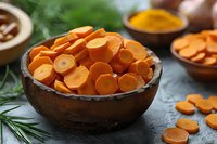 Fresh sliced carrots in rustic wooden bowl on dark background with herbs and spices, selective focus on orange vegetable pieces for cooking concept.