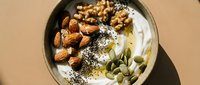 Overhead view of a bowl with yogurt, almonds, walnuts, pumpkin seeds, and poppy seeds on neutral background for healthy breakfast or wellness content.