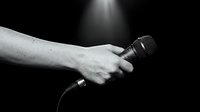 Hand holding microphone on dark stage with dramatic spotlight creating moody atmosphere for live performance, singing, karaoke or public speaking event.