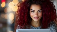 Young mixed-race woman with curly red hair smiling at camera in cozy indoor setting with warm bokeh lights background for lifestyle content.