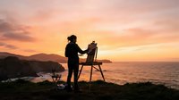 Silhouette of a woman standing on coastal cliff holding chair at golden hour sunset, ocean and mountains in background, outdoor leisure and contemplation.