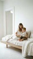 Young blonde mother reading book with toddler son on bed in bright minimalist bedroom creating cozy bonding moment together.