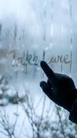 Hand in black glove writing Take Care on a foggy frozen window in winter, with snowy trees in the blurred background.