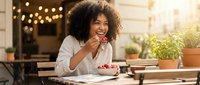 Young woman with curly dark hair and medium skin tone eating dessert at outdoor cafe table with plants, warm string lights, and notebook, lifestyle moment.