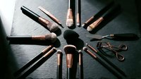 Professional makeup brushes and beauty tools arranged in circle on dark slate surface with rose gold accents for cosmetic application and styling routine.