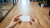 Hands holding blank white business card in modern office workspace with wooden floor and blurred background, suitable for branding and corporate identity projects.